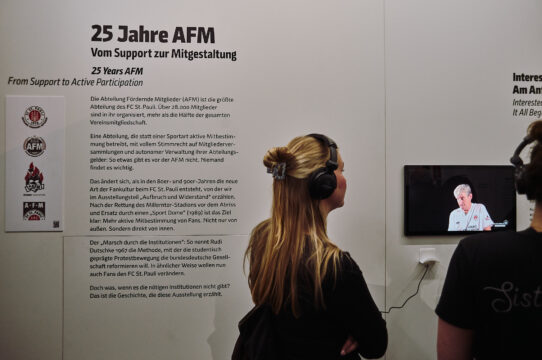 Zwei Besucher hören aufmerksam einer Präsentation über die 25-jährige Geschichte der AFM des FC St. Pauli zu.