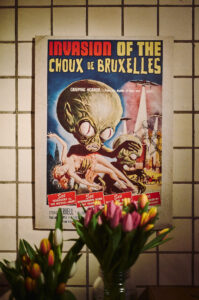 Ein farbenfrohes Retro-Poster von "Invasion of the Choux de Bruxelles" hängt über einer Vase mit frischen Tulpen