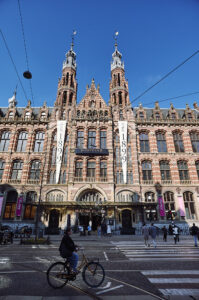Ein Radfahrer fährt vor der beeindruckenden Fassade der Magna Plaza in Amsterdam, die Shopping und Geschichte vereint.