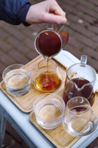 Eine Hand gießt aromatischen Kaffee aus einer Glaskaraffe in ein elegantes Gefäß auf einem Holztablett in Amsterdam.