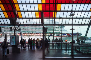 Menschen warten an der Bushaltestelle in Amsterdam, während ein großes Kreuzfahrtschiff hinter einer bunten Glasdecke sichtbar ist.